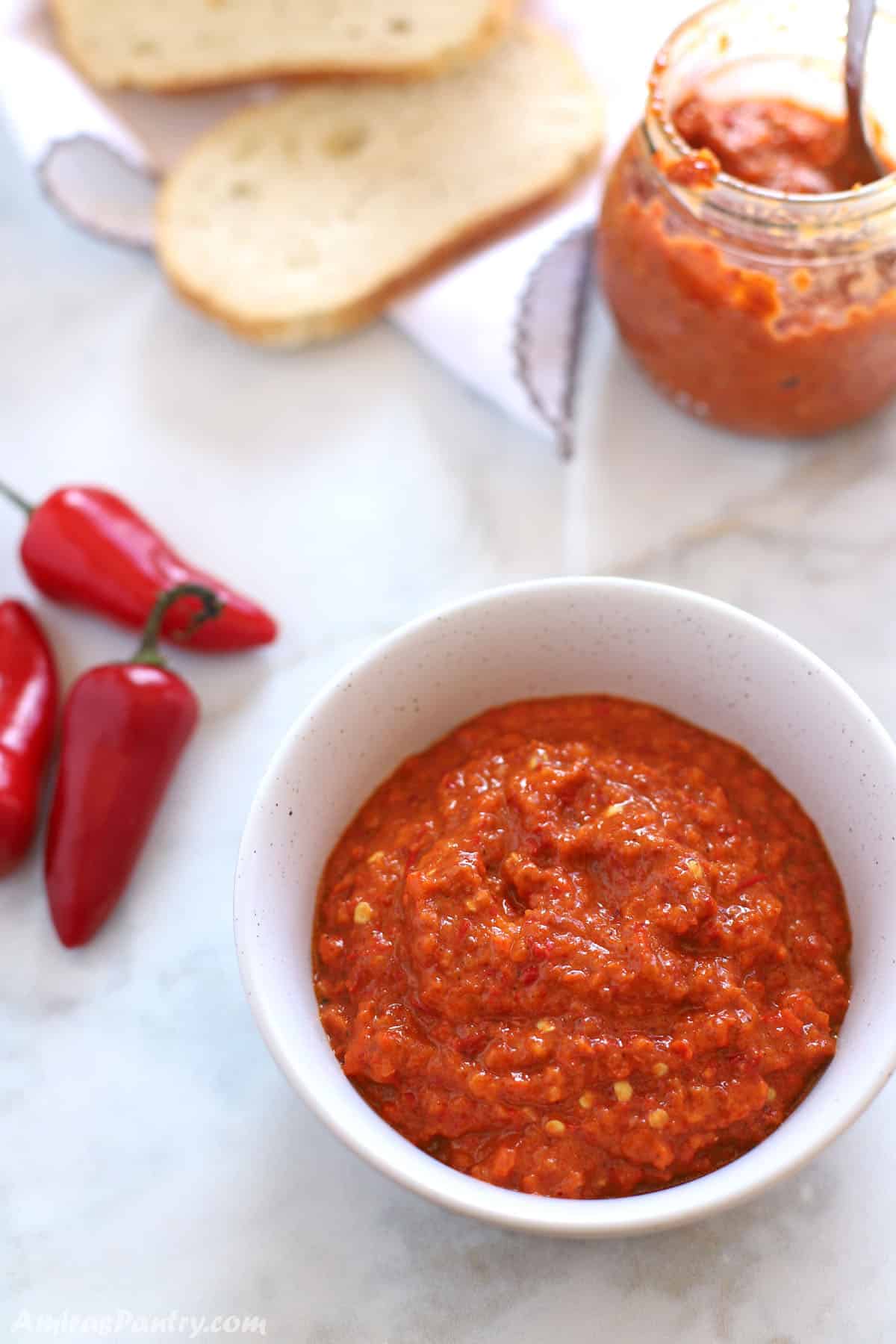 Harissa in a white bowl with a jar half empty in the back, bread and some red peppers all placed on a marble white table.