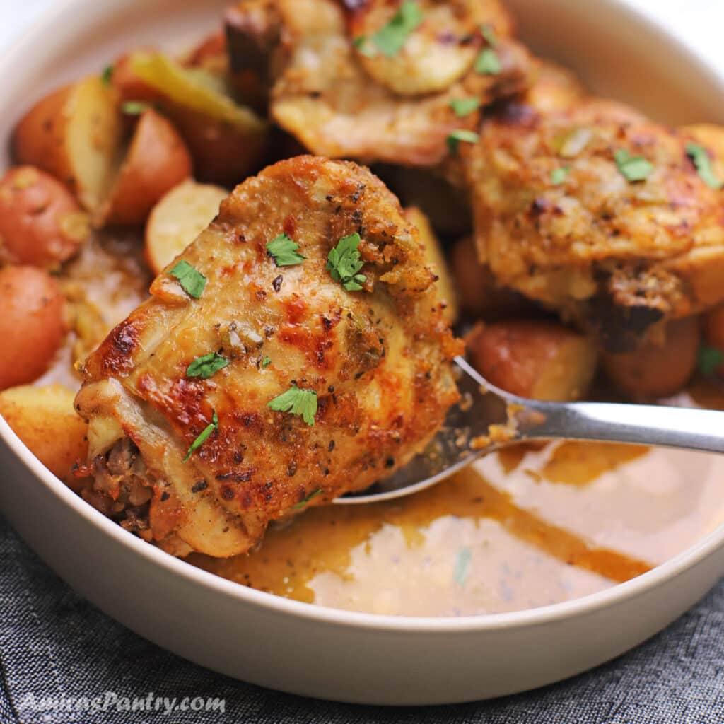 A spoon scooping chicken thighs from a platter with potatoes on the back.