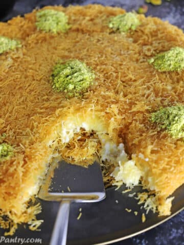 Knafeh with a piece cut out of it andgarnished with crushed pistachios on a black metal platter..