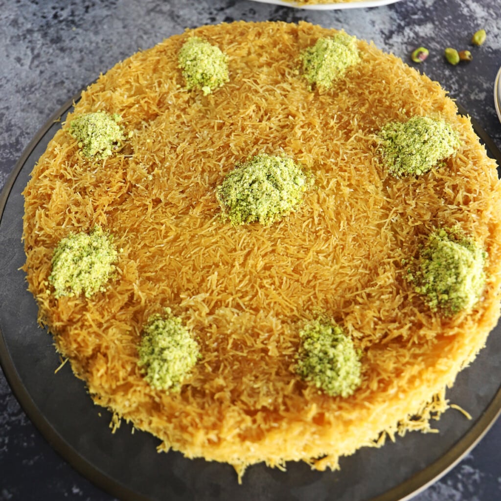 Knafeh garnished with crushed pistachios on a black metal platter.