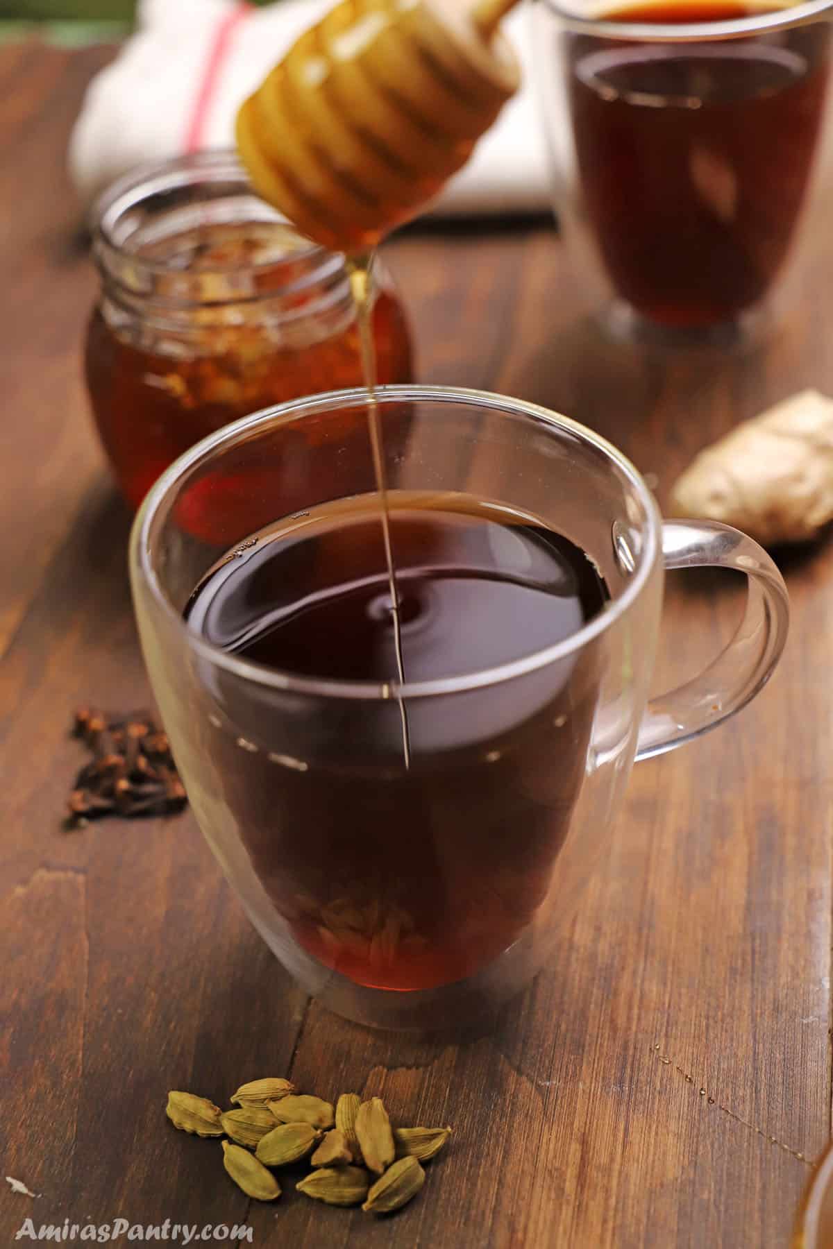 A honey spoon pouring honey into a cup of cardamom tea.