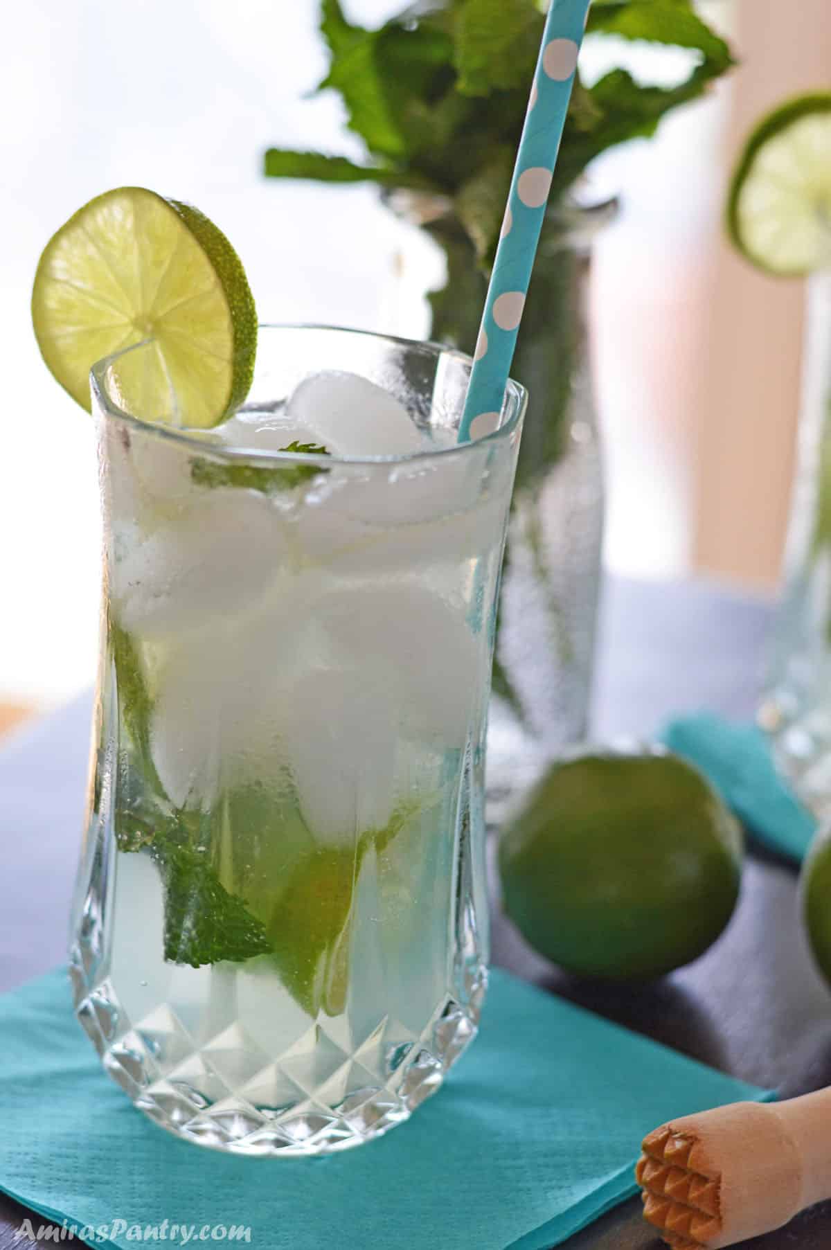 A tall glass cup of virgin mojito on a light blue napkin with lime and mint leaves on the back.