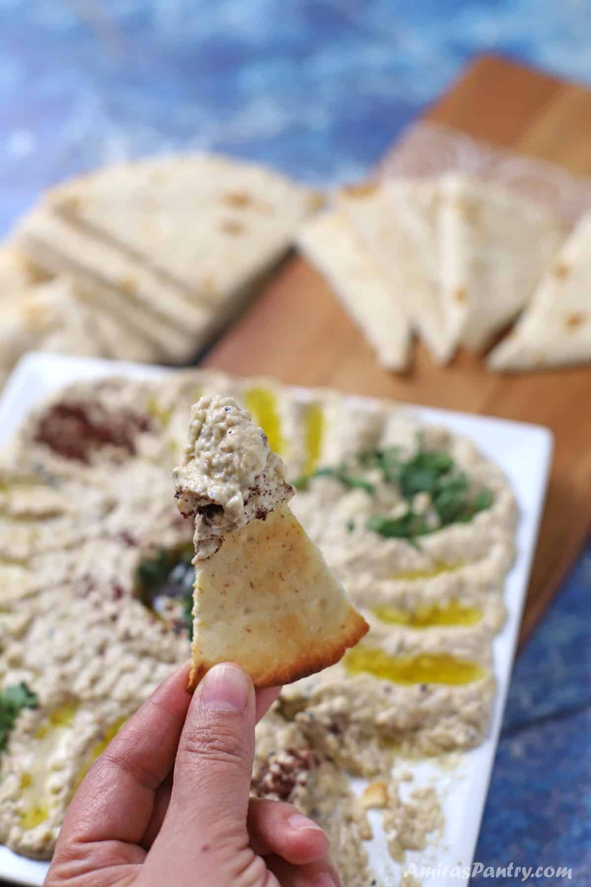 A hand holding a piece of pita wedge dipped in mutabbal.