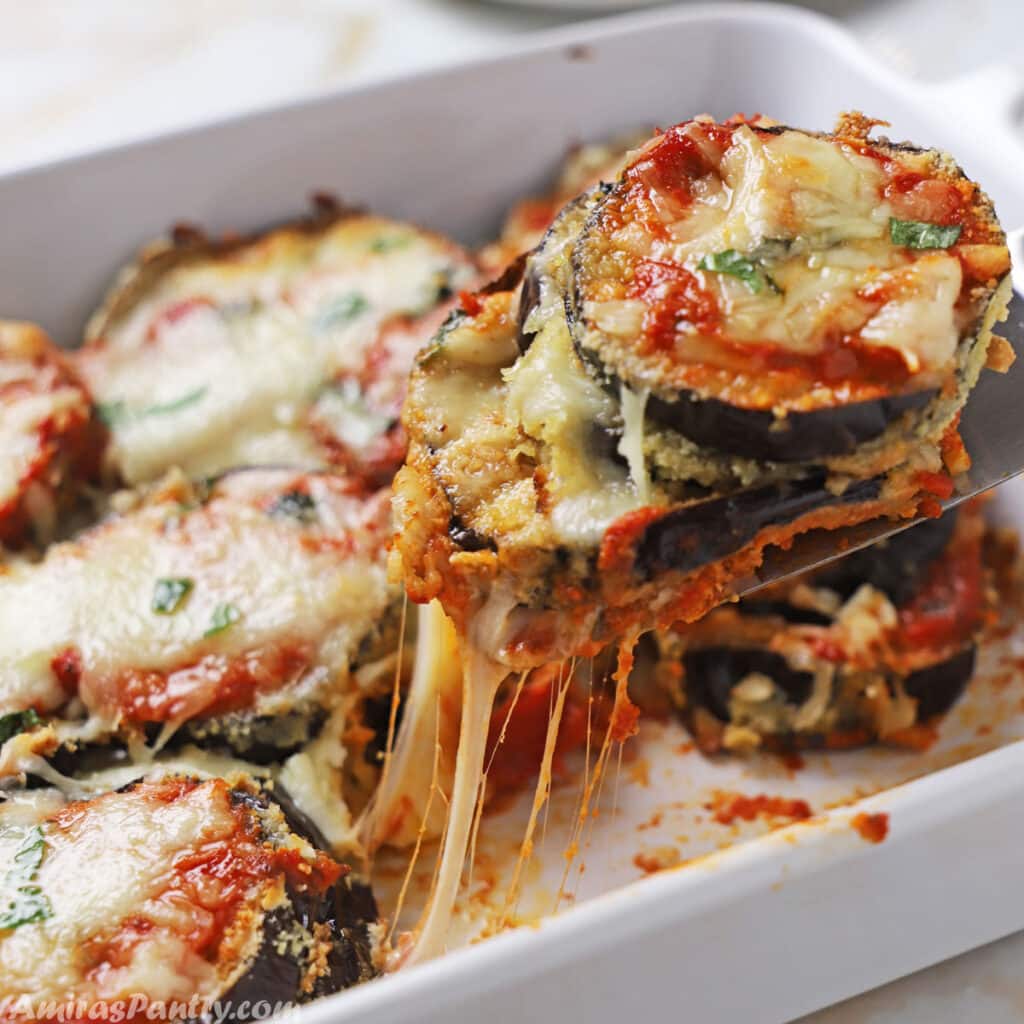 A hand scooping some eggplant parmesan out of a white deep casserole.