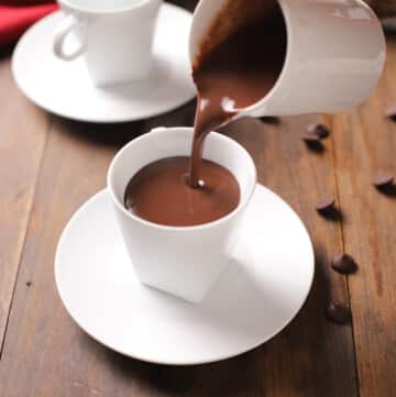 A white carafe pouring hot chocolate into a white mug and all placed on a wooden surface.