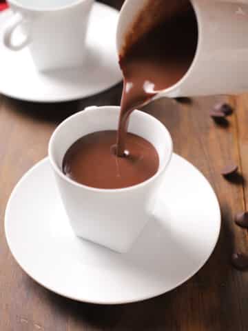 A white carafe pouring hot chocolate into a white mug and all placed on a wooden surface.