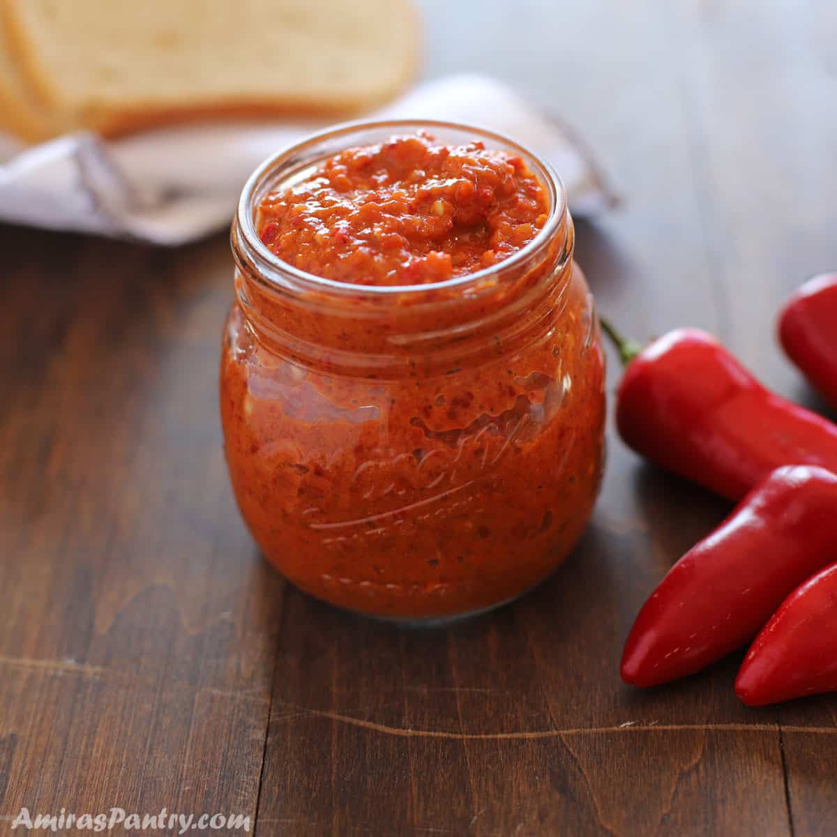 A jar of Harissa on a wooden surafce with some chili peppers on the side and bread on the back.
