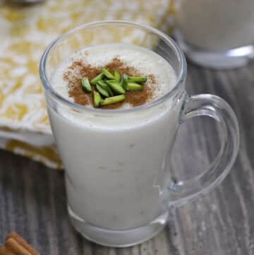 A glass cup of sahlab on a wooden countertop garnished with cinnamon and pistachios.