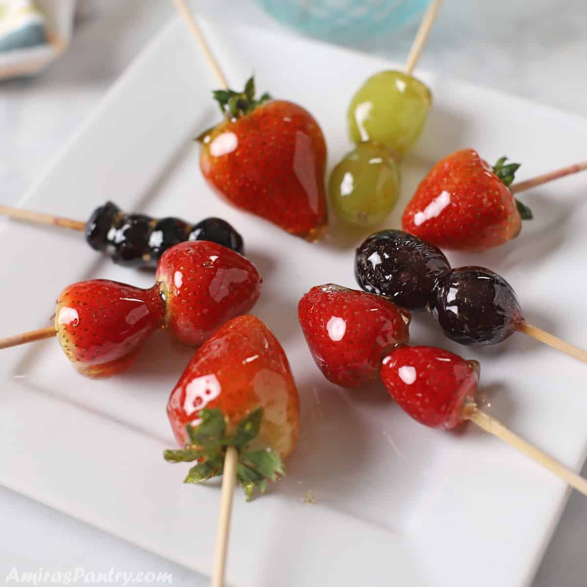 Strawberries Tanghulu Skewers  on a white plate with grapes and blue berries tanugul as well.