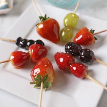 Strawberries Tanghulu Skewers on a white plate with grapes and blue berries tanugul as well.