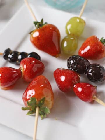 Strawberries Tanghulu Skewers on a white plate with grapes and blue berries tanugul as well.