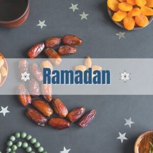 Dates shaped like a crescent placed on a dark table with bowls of almonds and Turkish dried apricot and a green prayer beads.