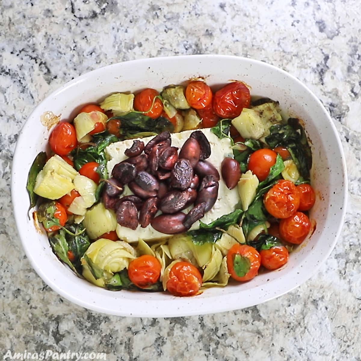 A white dish with baked feta topped with kalamata olives and other vegetables on the dish.