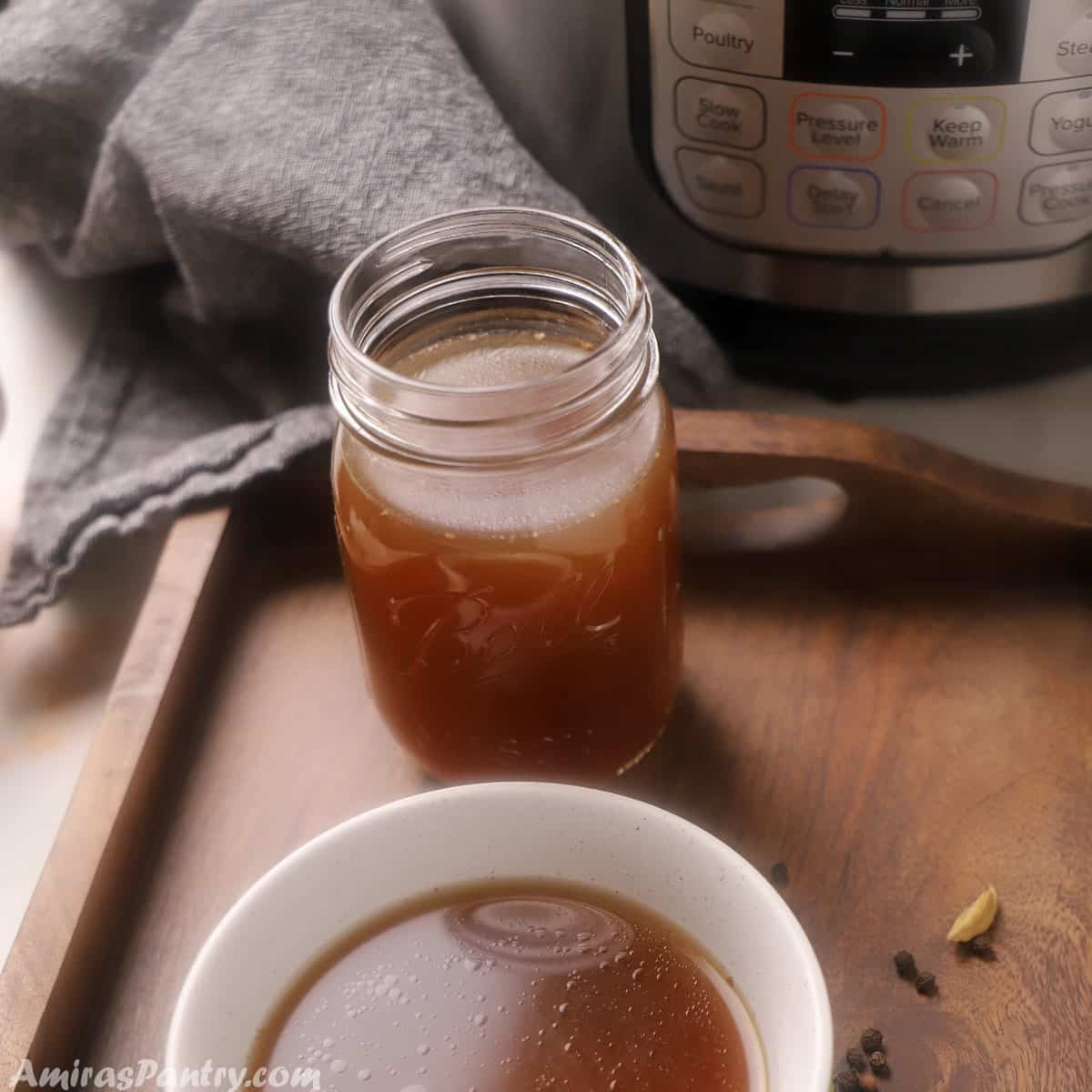 A mason jar filled with bone broth and a bowl with bone broth placed infront if an instant pot on a wooden tray.