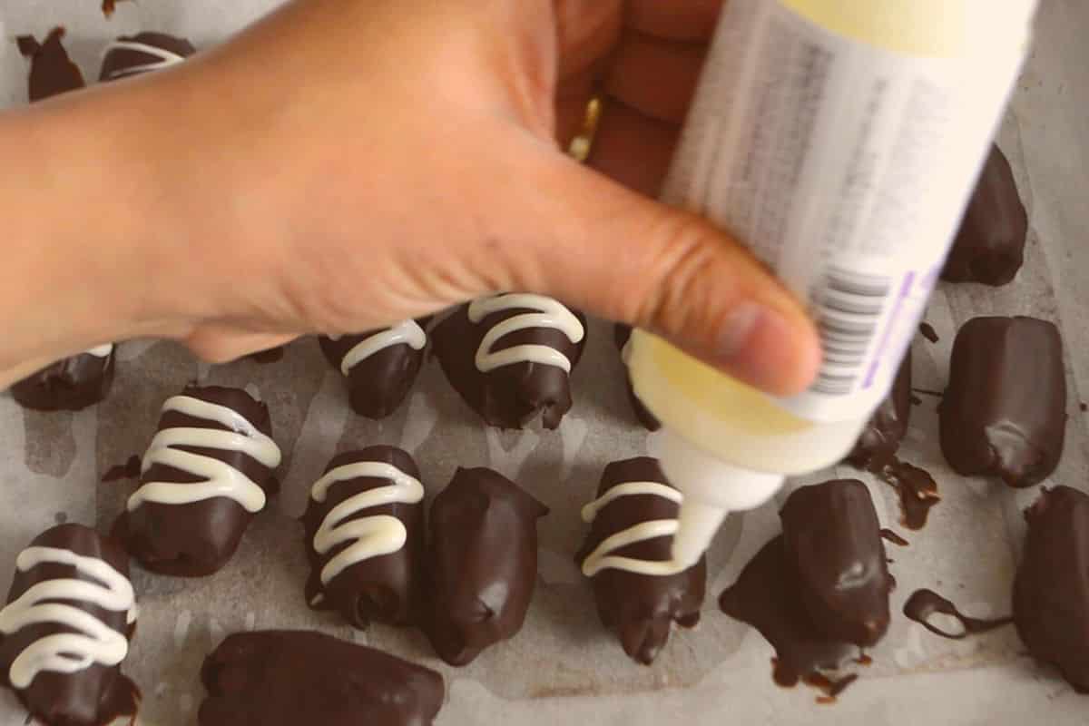 A hand holding a decorating tube to decorate the chocolate covered dates with white chocolate.