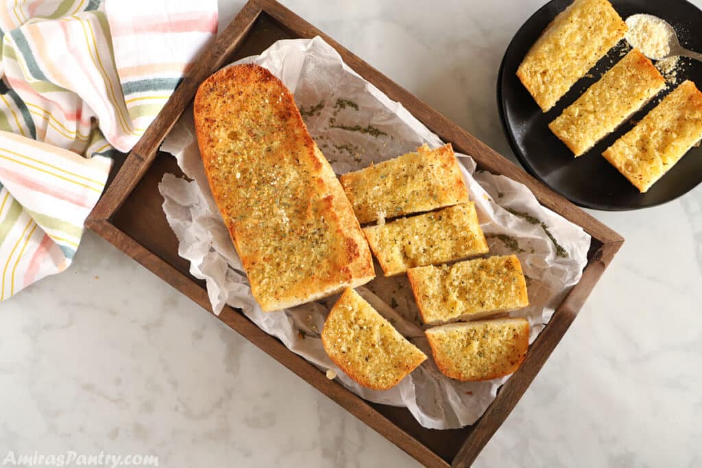 A top view image of garlic bread placed on a wooden tray with a plate of sliced garlic bread on the side.