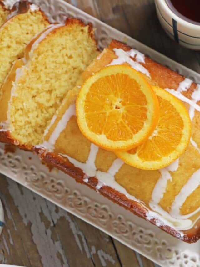 A top view of a sliced orange cake in a white platter with a cup of tea on the side.