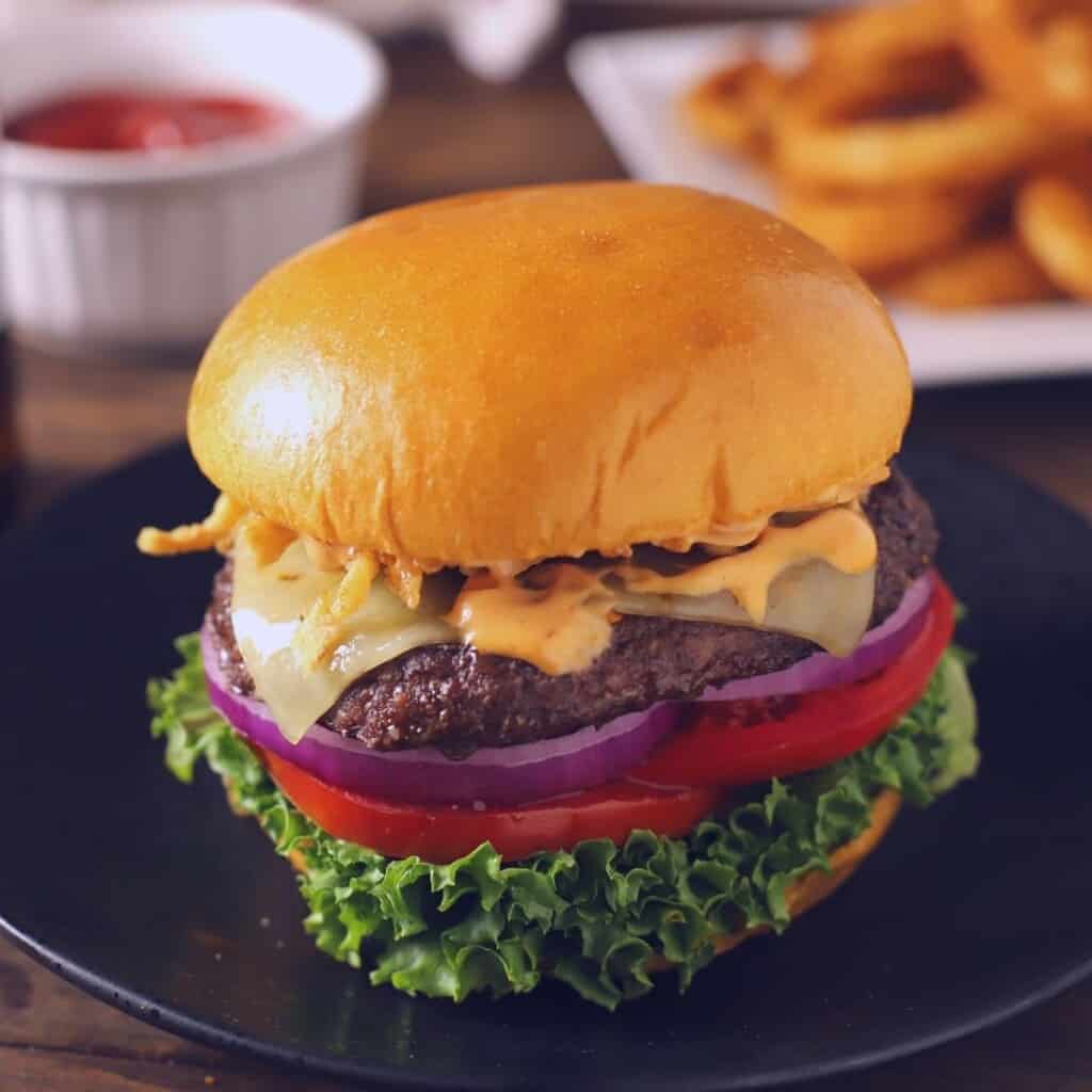 Burger sandwich on a wooden surface with sauces and onion rings on the back.