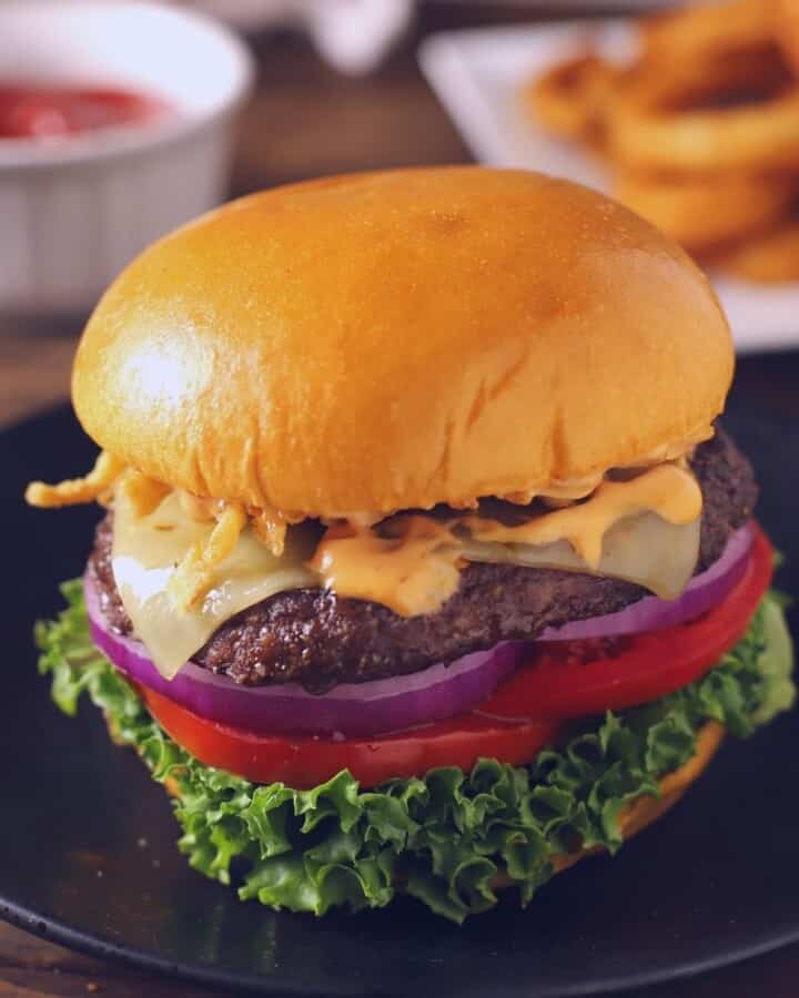 Burger sandwich on a wooden surface with sauces and onion rings on the back.