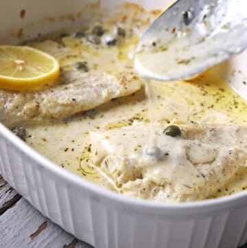 A spoon scooping some creamy sauce over baked fish lemon in a white pan.