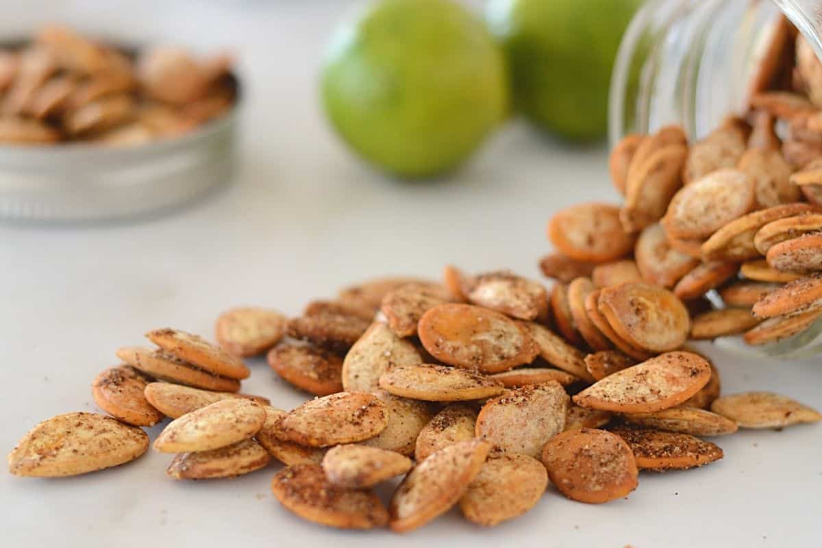 Spicy roasted pumpkin seeds falling from a glass jar on a white marble surface.