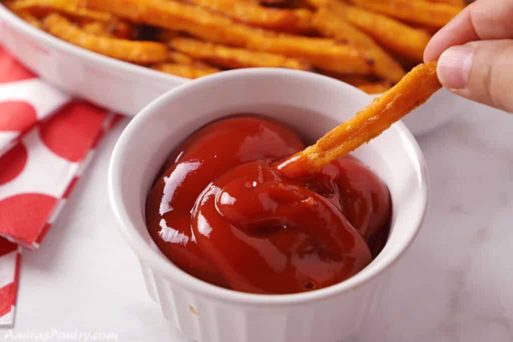 A hand dipping sweet potato fries in a small plate of ketchup.