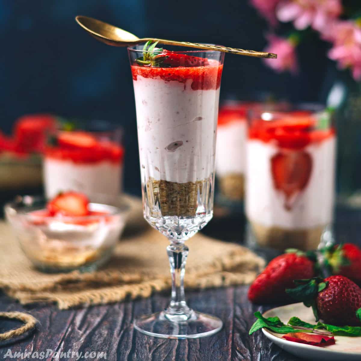 A tall glass of strawberry cheese cake with a golden spoon resting on its top.