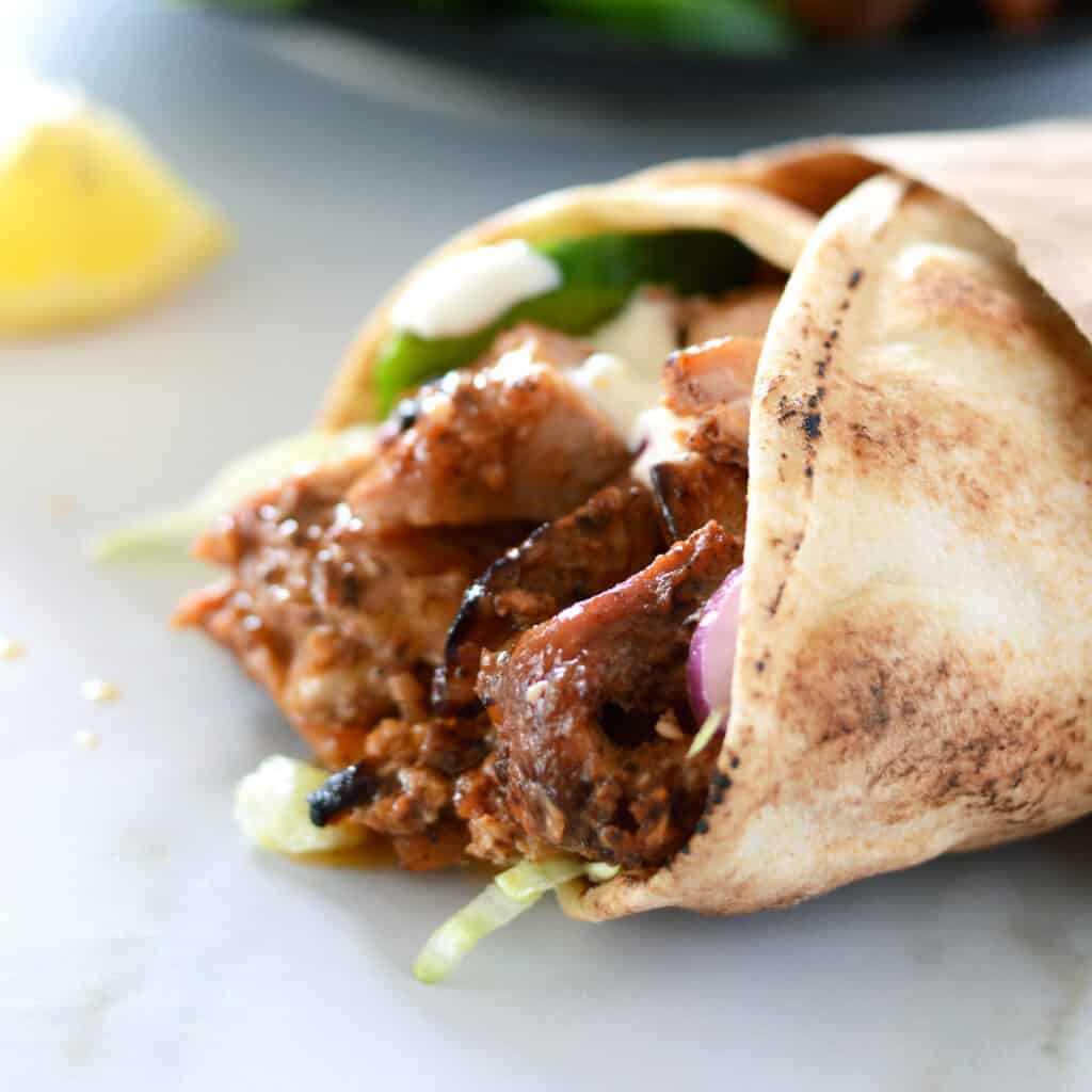 A close up picture of a chicken shawarma wrap placed on a awhite countertop with a lemon wedge on the side.