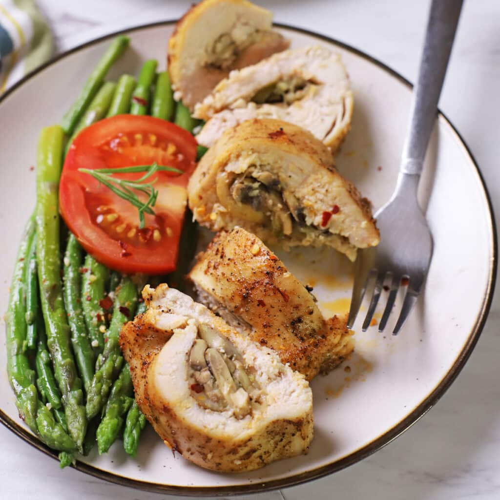 Air fryer stuffed chicken breas cut into pieces on a white plate with a fork, asparagus and tomato slice in the plate.