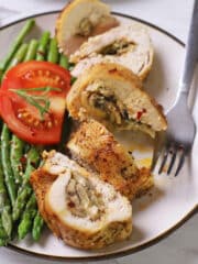 Air fryer stuffed chicken breas cut into pieces on a white plate with a fork, asparagus and tomato slice in the plate.