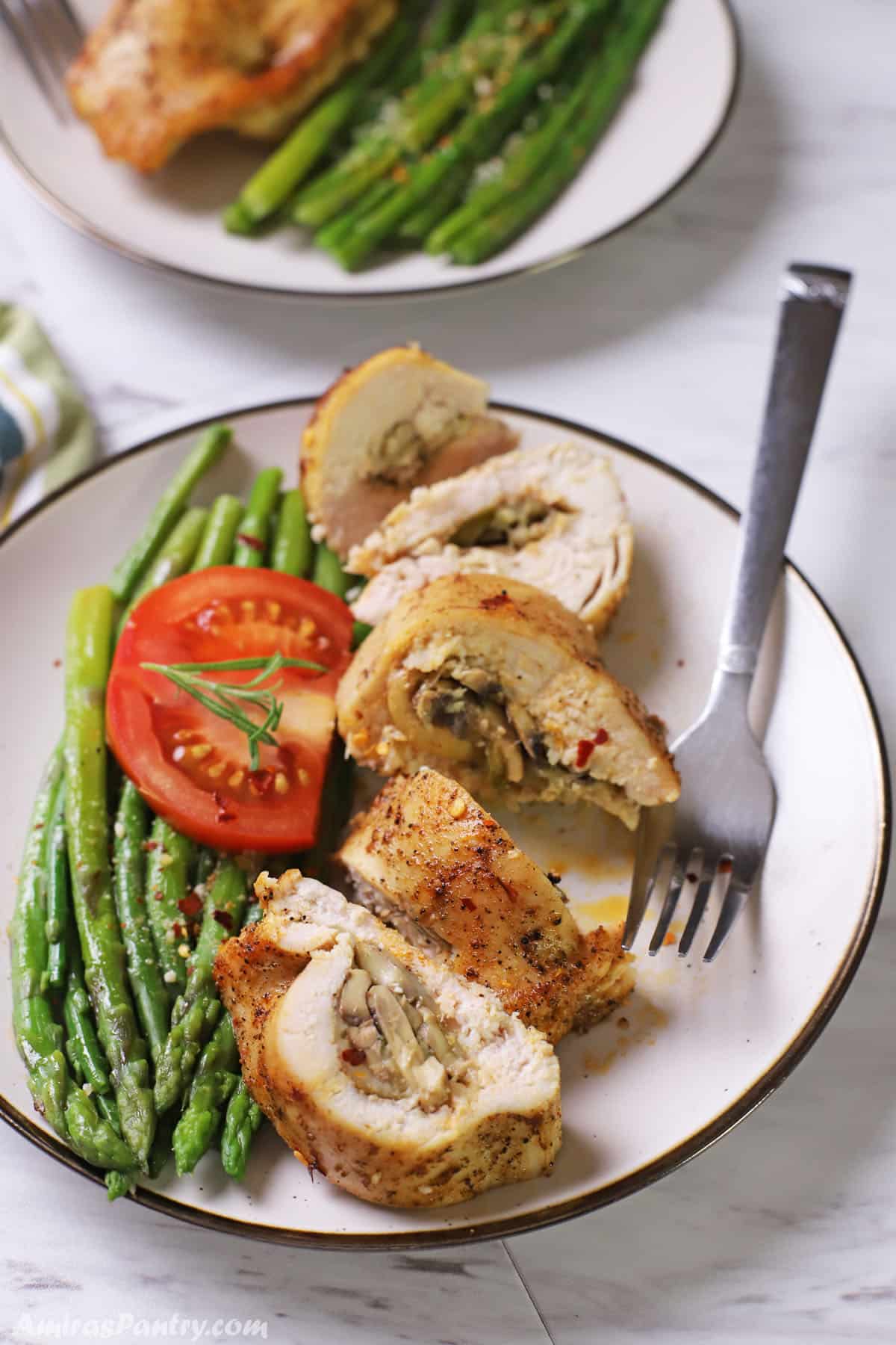 A white plate with stuffed chicken breast cut into pieces with some asparagus and tomato in the plate as well.