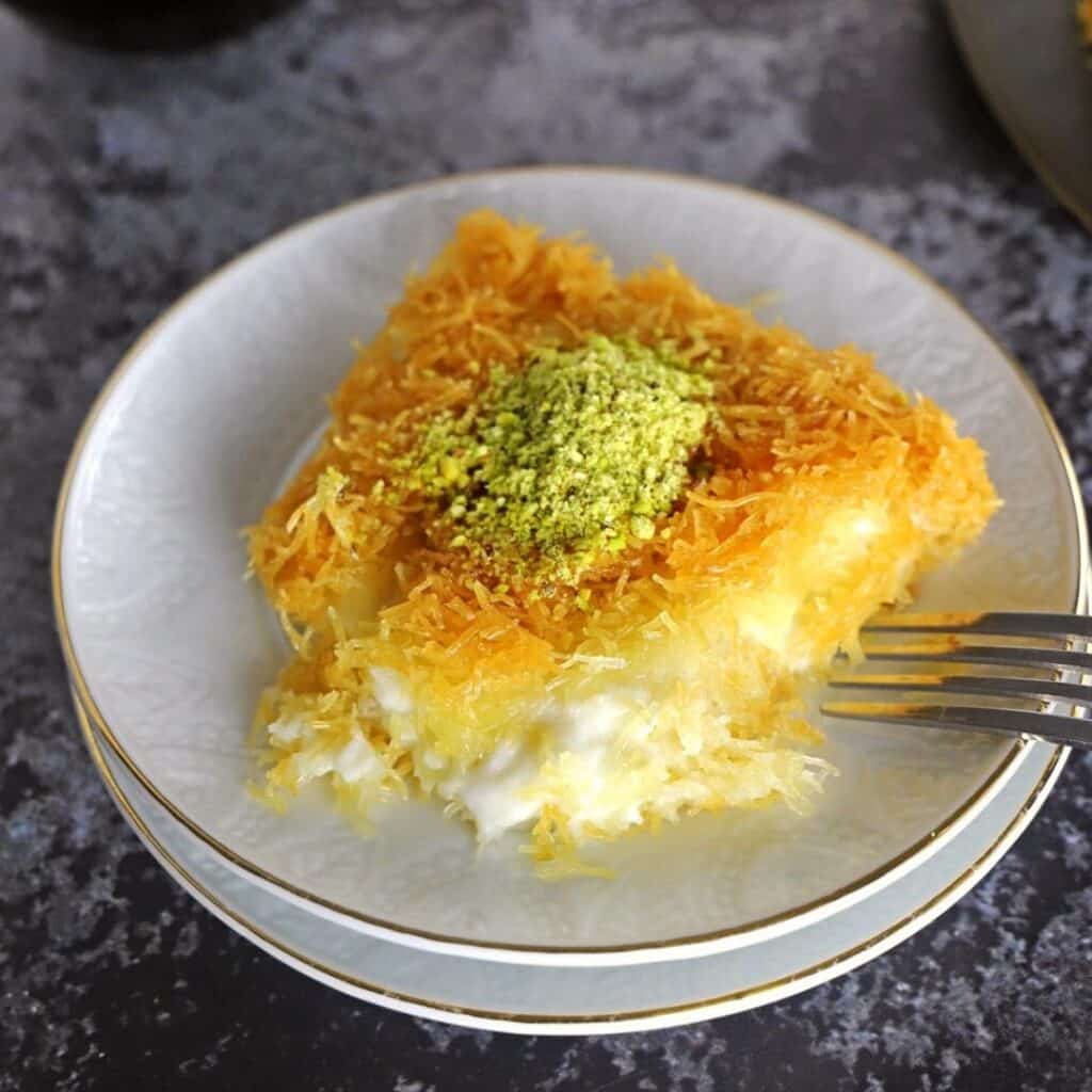 A piece of kunafa on a white dessert plate with a fork next to it.