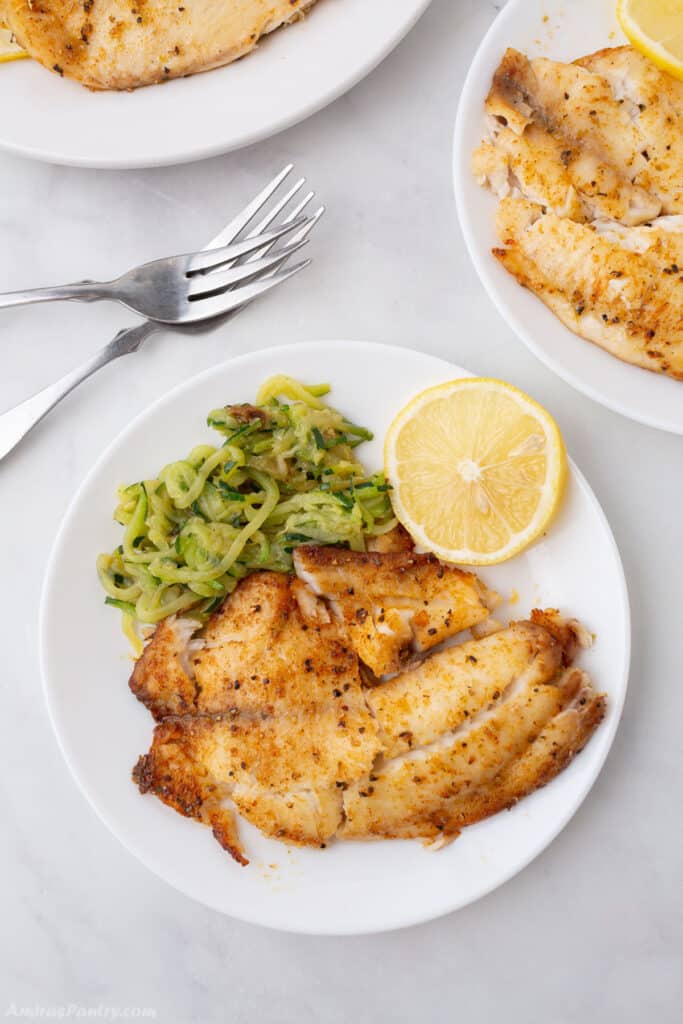 A bird's eye view of a plate with tilapia and some veggies and forks on the side.