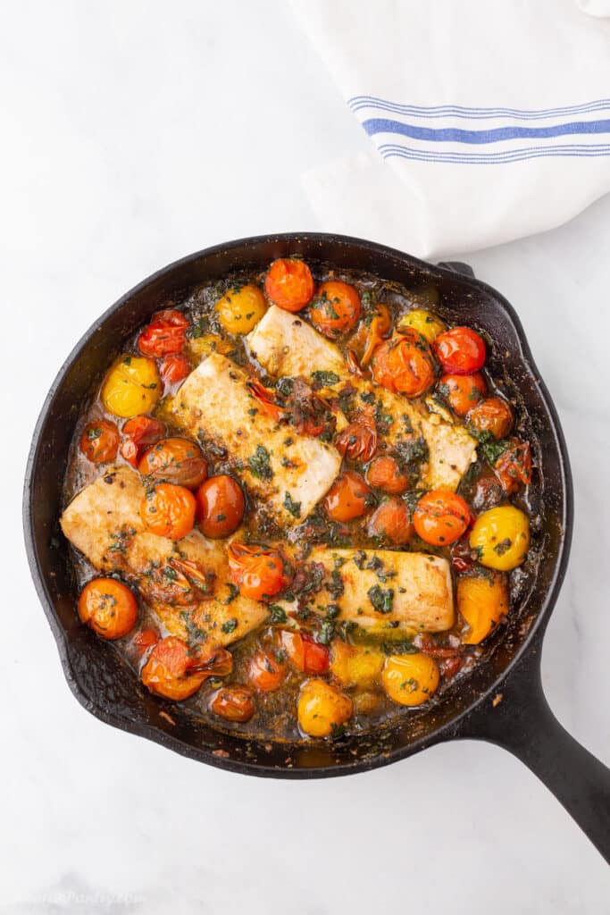 A bird's eye view of a cast iron skillet with baked fish and grape tomatoes.
