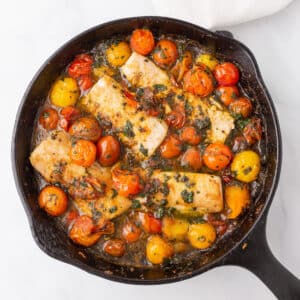 A bird's eye view of a cast iron skillet with baked fish and grape tomatoes.