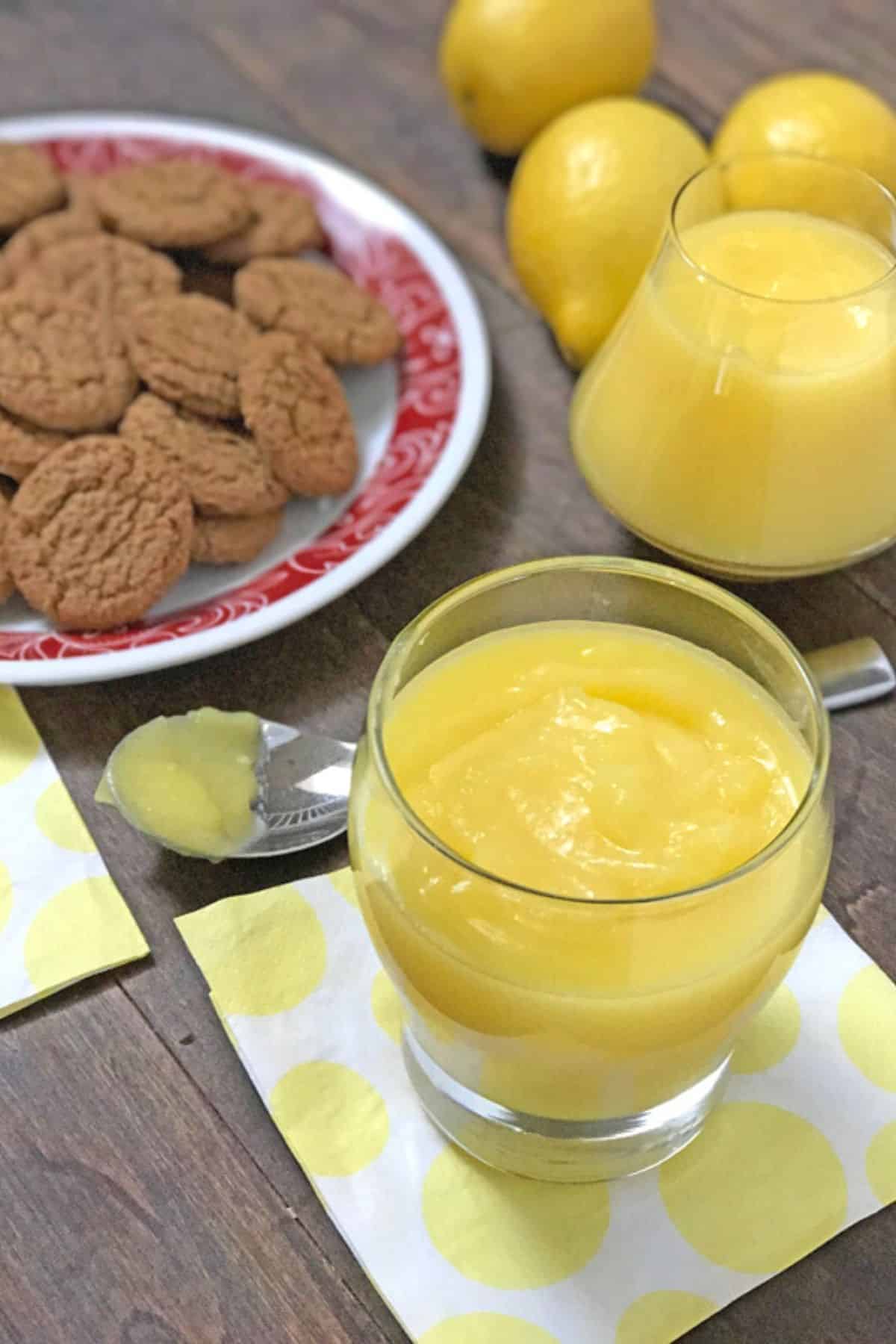Two jars of lemon curd with lemon and cookies on a wooden table.