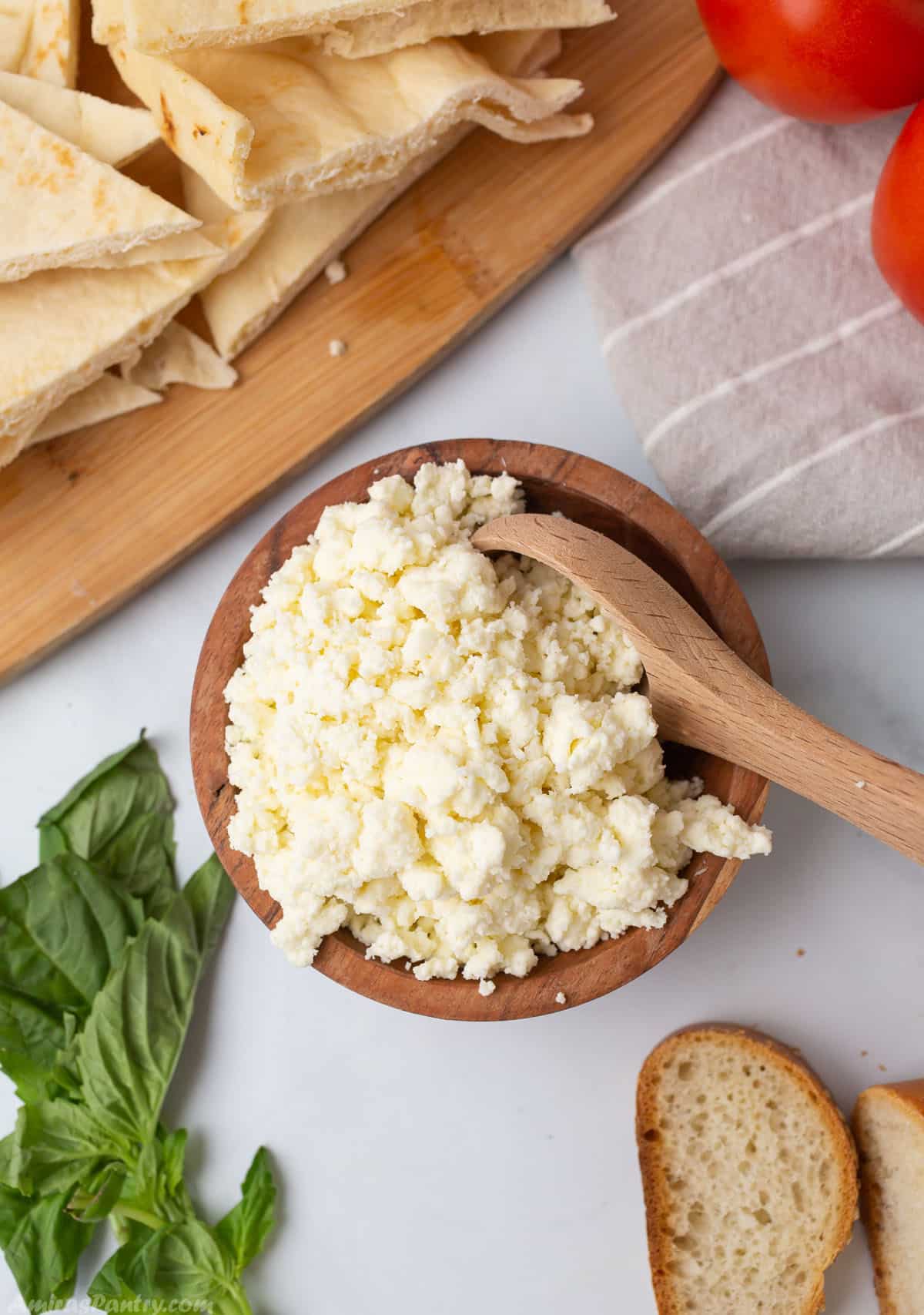 Ricotta cheese on a wooden bowl with a wooden spoon, some bread and basil on the picture.