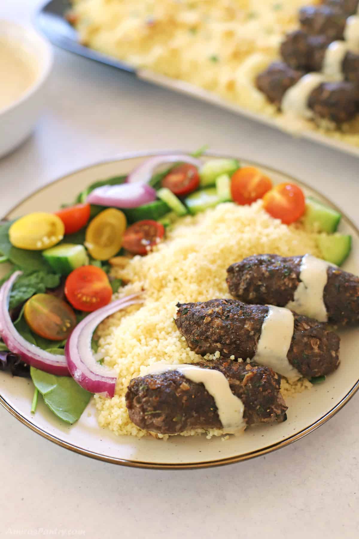 Kafta in a white plate drizzled with tahini sauce with green salad and couscous on the side.