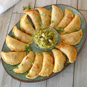 A green platter with empanadas and a small bowl of guacamole.
