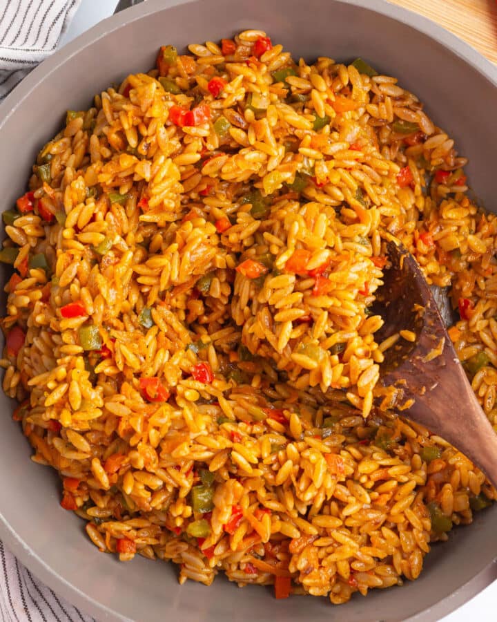 A large skillet of orzo with vegetables and a wooden spoon in it.