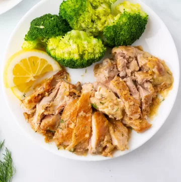 Lemon dill chicken on a white plate with broccoli and a slice of lemon.