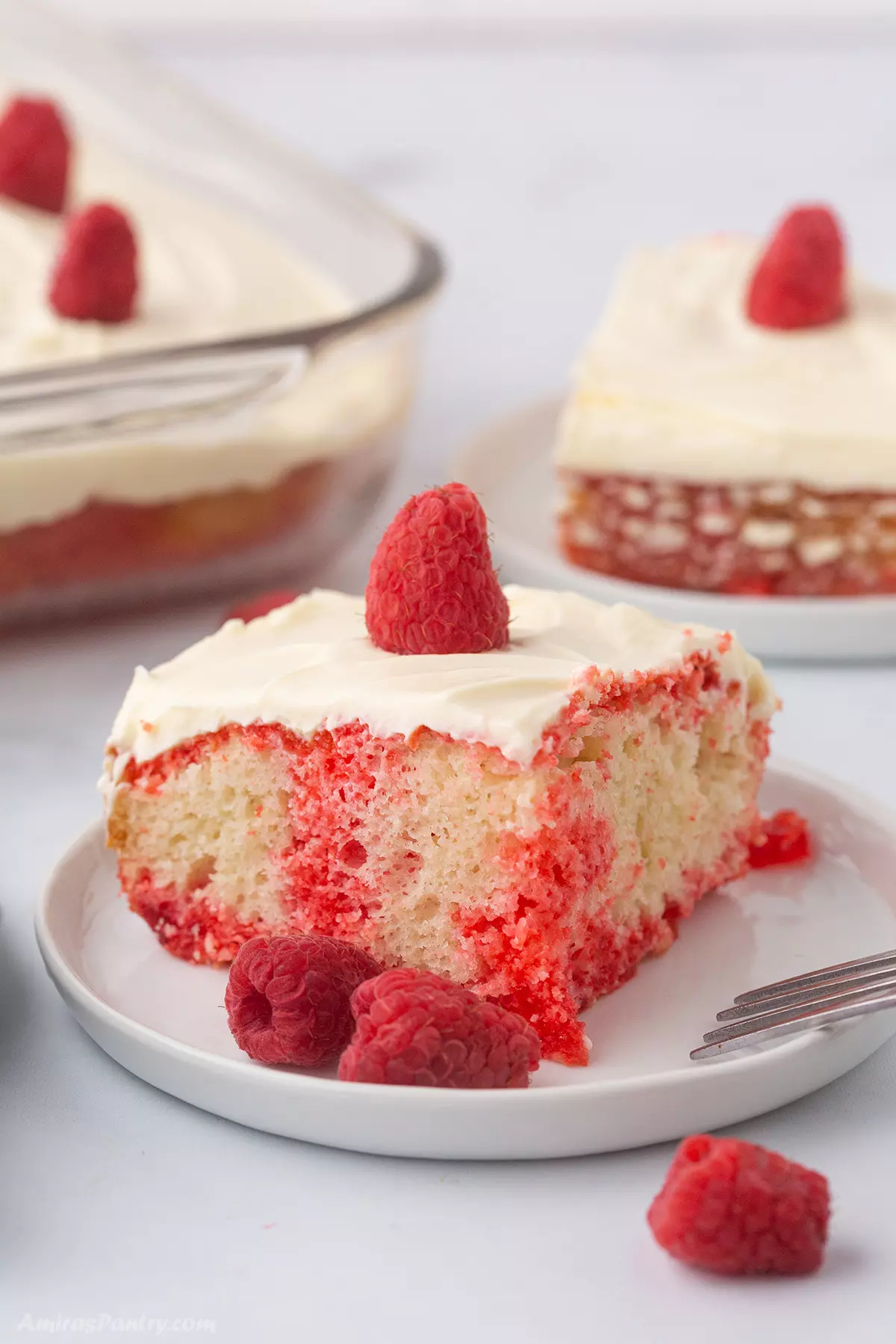 A slice of raspberry cake on a white dessert plate and fresh raspberries.