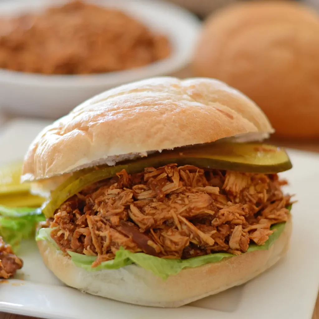 a pulled bbq chicken sandwich on a white plate with a bowl of pulled chicken in the back