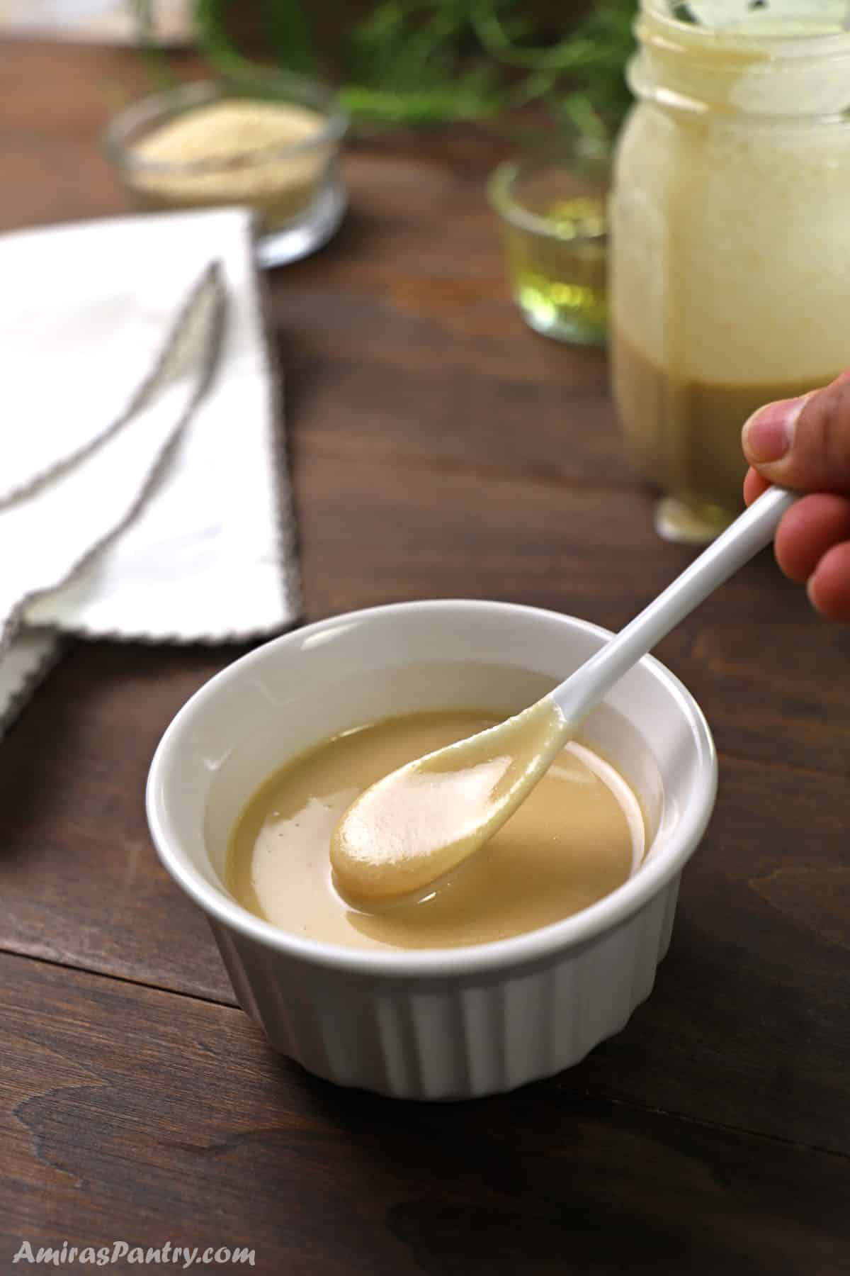 A spoon scooping some freshly made tahini paste out of a small bowl with a jar of tahini in the back.