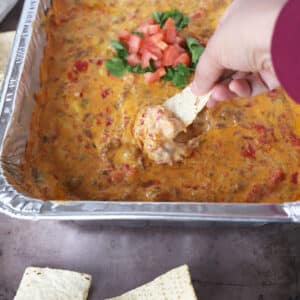 A hand scooping some smoked queso dip out of a tin foil casserole.