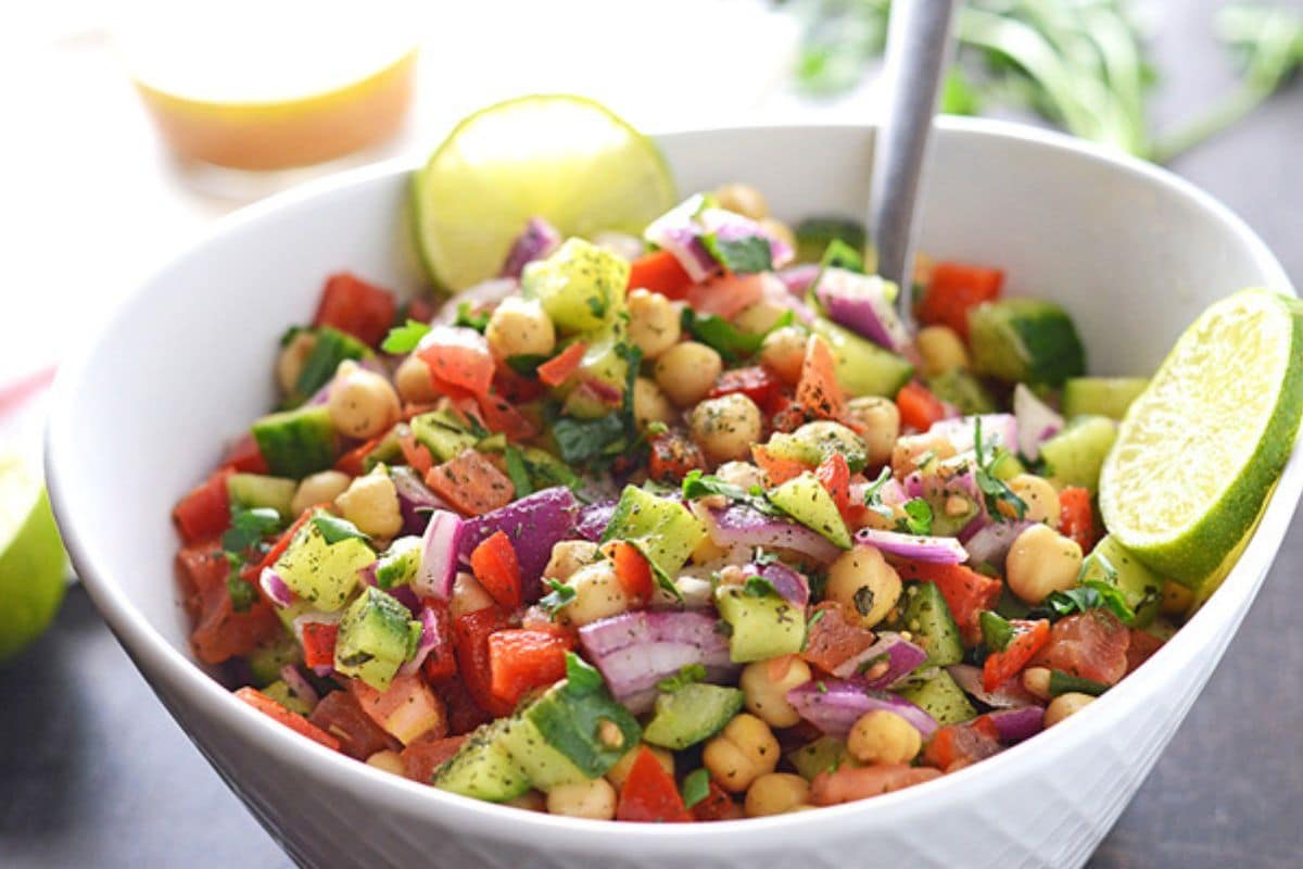 A white bowl of chickpea salad with a spoon and lemon wedges.