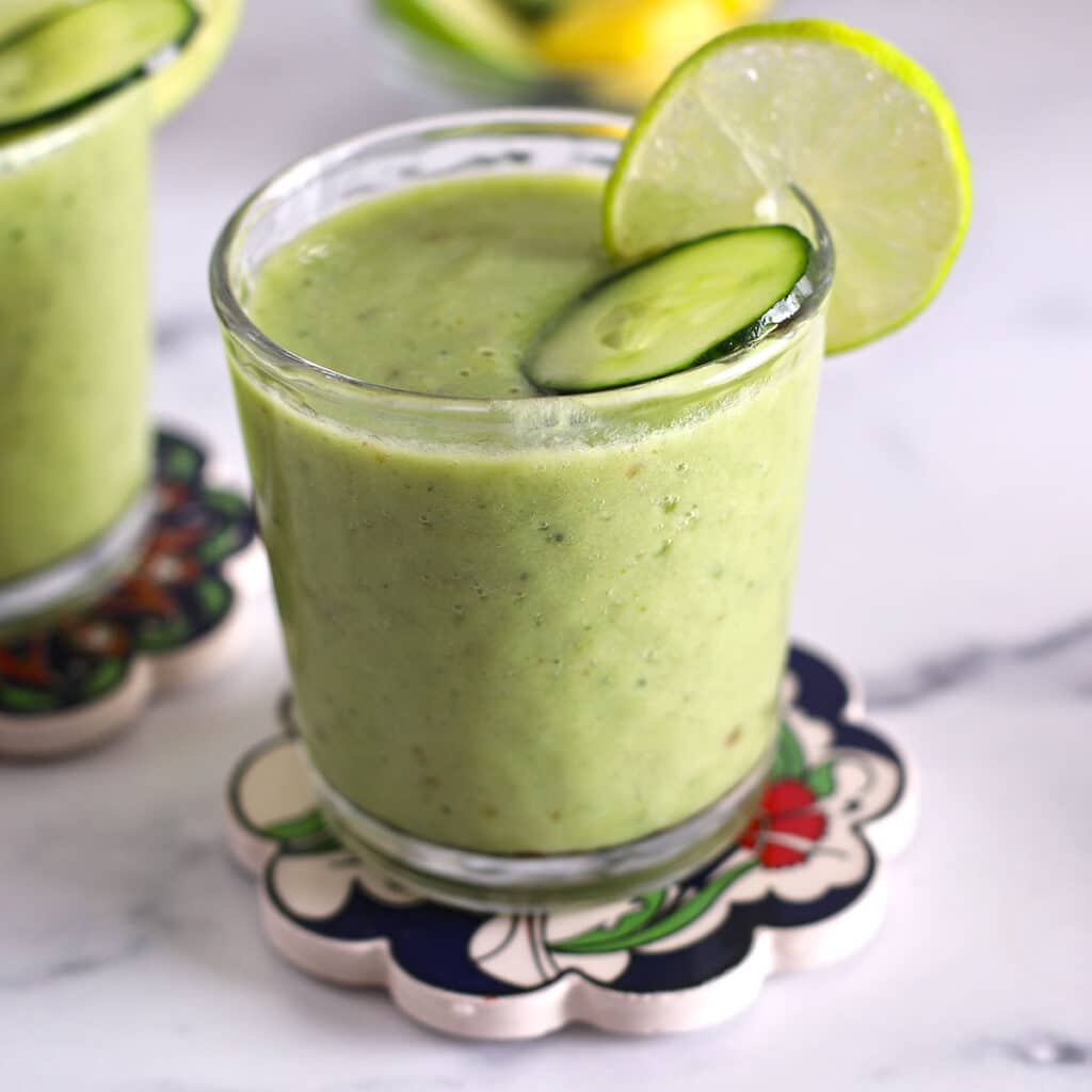 A glass cup with green pineapple cucumber smoothie with lime and cucumber slices.