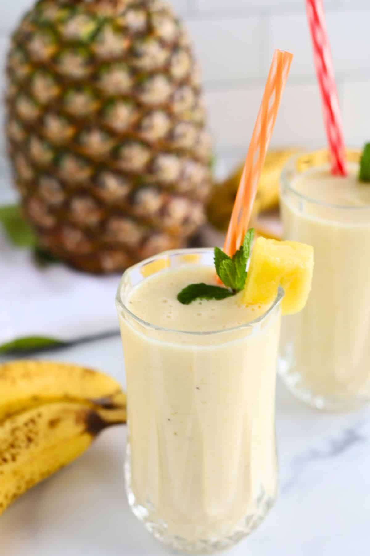 Glasses of banana pineapple smoothie with pineapple and bananas at the back.