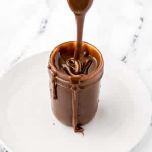 A spoon drizzling some hot fudge sauce in a mason jar.