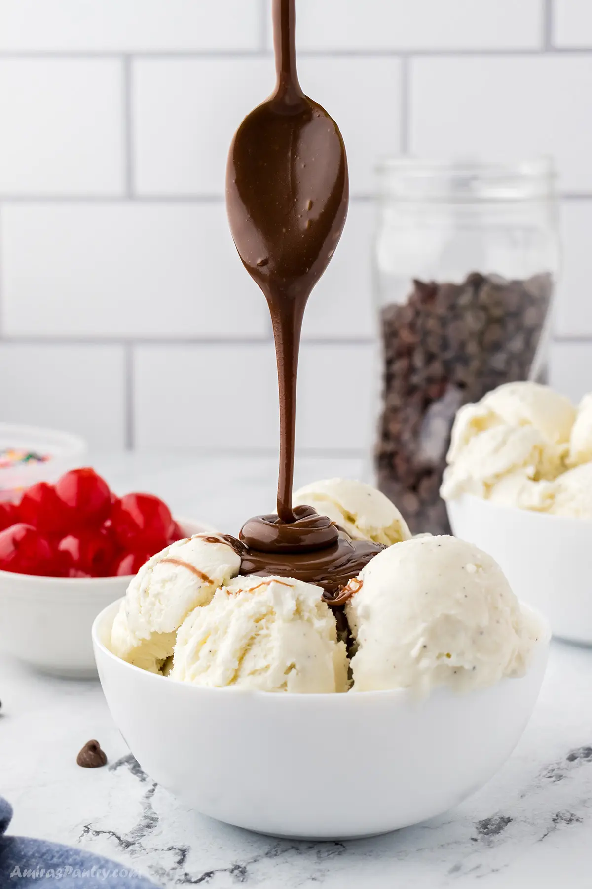 A spoon drizzling some hot fudge sauce over a bowl of ice cream.